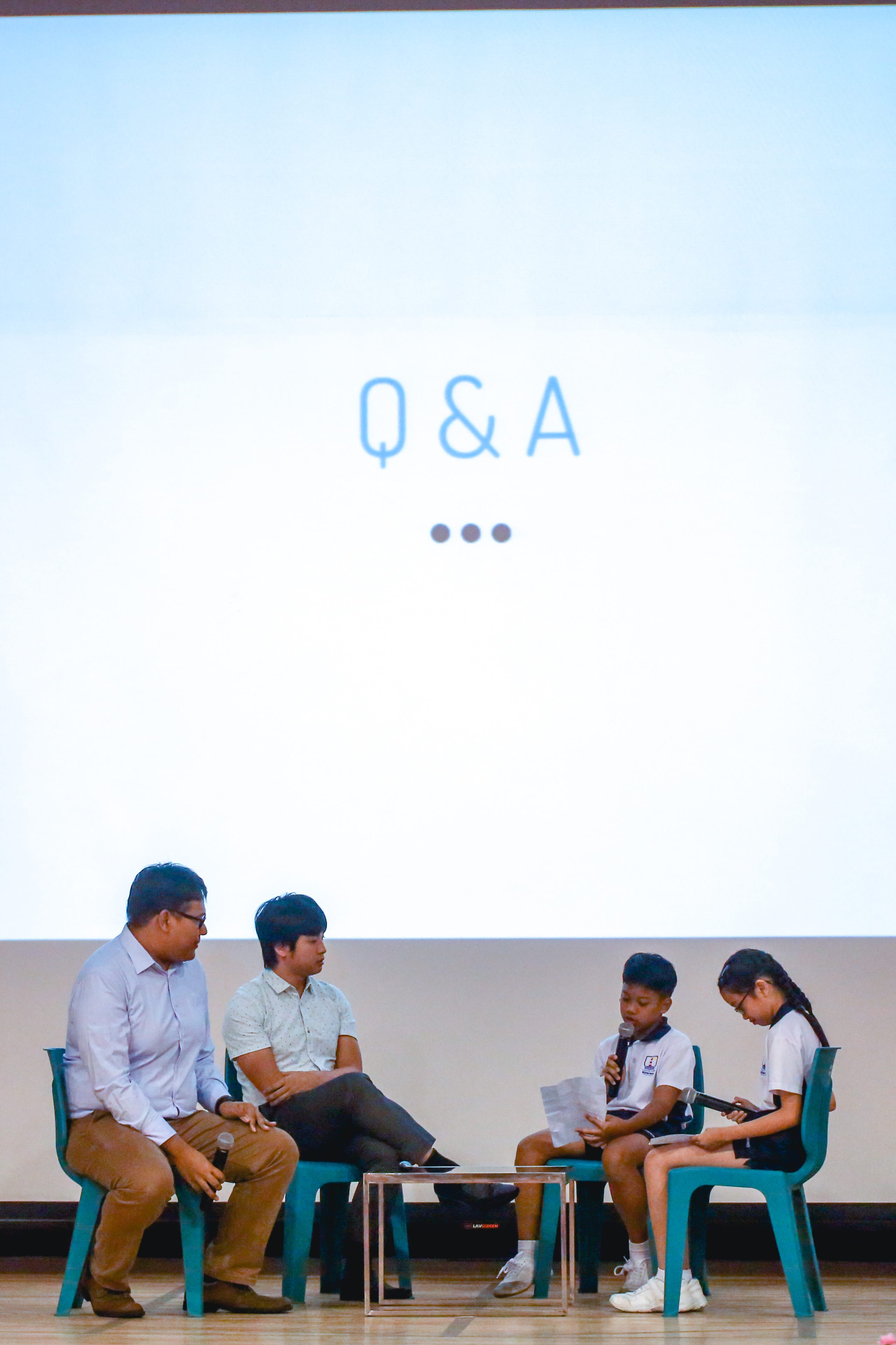 Four people sit on teal chairs on a stage during a Q&A session, with two adults and two children holding microphones.